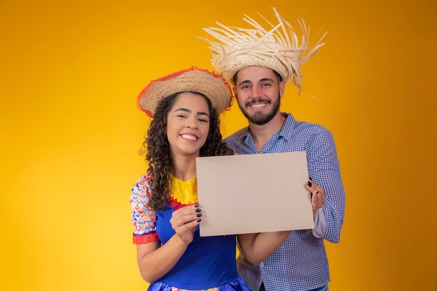 Pareja brasileña con ropa tradicional para Festa Junina sosteniendo un cartel con espacio para copiar