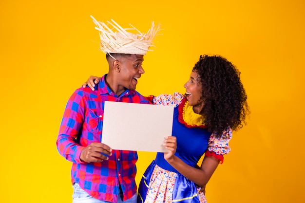 Pareja brasileña con ropa tradicional para Festa Junina sosteniendo un cartel con espacio para copiar