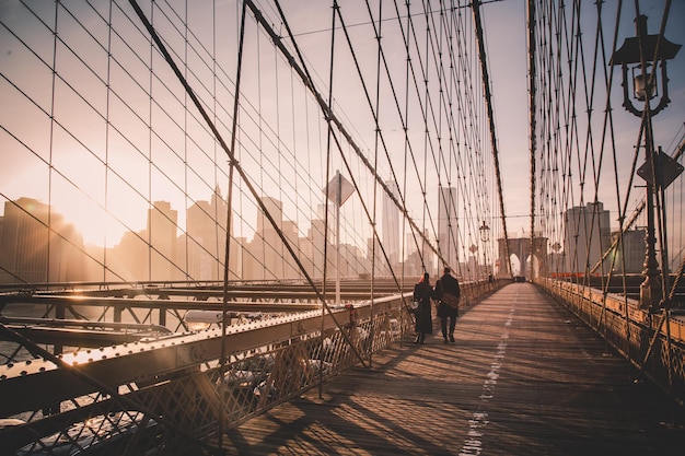 Pareja caminando por el sendero peatonal a través del puente de Brooklyn. Horizonte del centro de Manhattan de Nueva York en la puesta del sol con rascacielos iluminados sobre el panorama del East River como se ve desde el puente de Brooklyn.