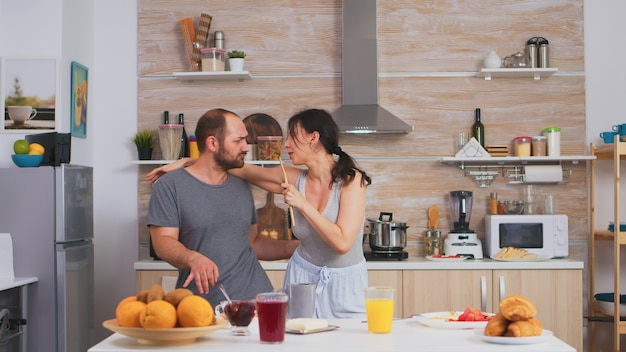 Pareja cantando en la cocina por la mañana en pijama durante el desayuno. Esposa y esposo despreocupados riendo divirtiéndose divertido disfrutando de la vida auténtica gente casada relación feliz positiva