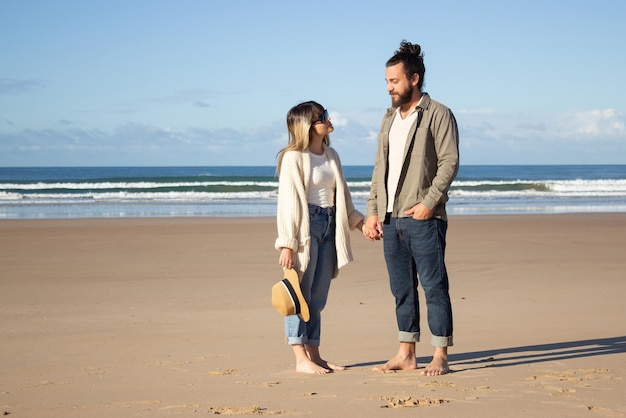 Pareja cariñosa caminando por la playa. Hombre y mujer barbudos con gafas mirándose, tomándose de la mano. Amor, vacaciones, concepto de afecto.