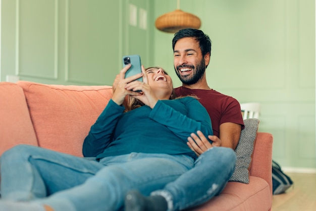 Pareja en casa a la luz del día tumbada en el sofá relajándose y uniéndose sosteniendo el teléfono móvil