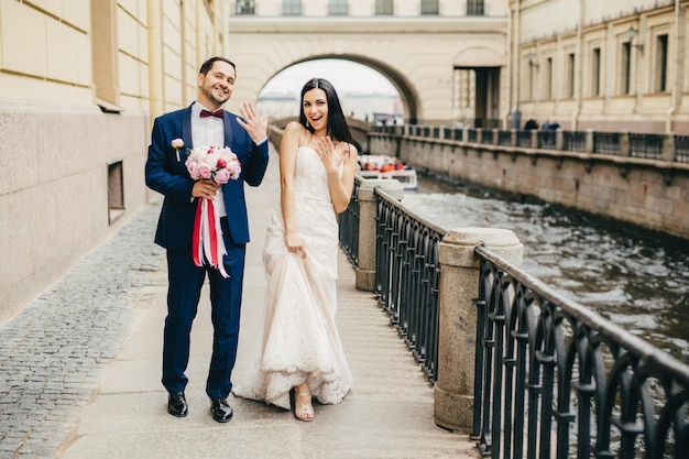 Pareja casada emocionada muestra anillos en los dedos camina en poses de puente en la cámara para hacer fotos inolvidables Mujer morena con vestido blanco y hombre guapo sostiene anillos de exhibición de ramo de novias