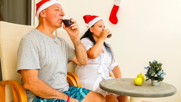 Una pareja casada con gorros de Papá Noel celebra la Navidad en el balcón en una mesa con un pequeño árbol de Navidad y un cóctel.