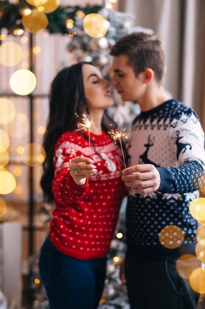 Pareja casada joven y atractiva en el fondo de un interior de Año Nuevo y un árbol de Navidad decorado besándose y sosteniendo luces de bengala