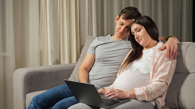 Una pareja casada que espera un bebé con cómodos atuendos domésticos hace compras en línea para un futuro hijo usando una computadora portátil y abrazándose en el sofá en la sala de estar