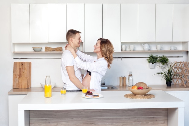 Pareja de casados jóvenes abraza de pie junto a la mesa en la cocina. El esposo abraza a su esposa embarazada, poniendo sus manos sobre su gran barriga, los padres se miran el uno al otro. Estilo de vida, pareja enamorada, gente feliz