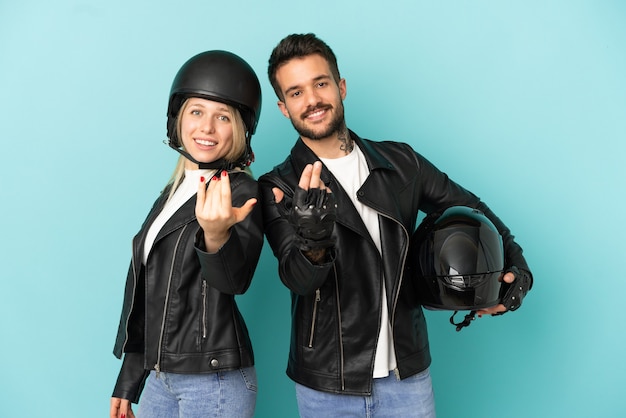 Pareja con casco de moto sobre fondo azul aislado que invita a venir con la mano. Feliz de que hayas venido