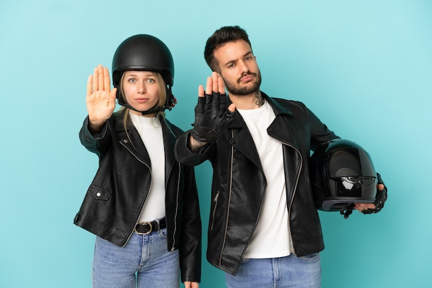 Pareja con casco de motocicleta sobre fondo azul aislado haciendo gesto de parada negando una situación que piensa mal