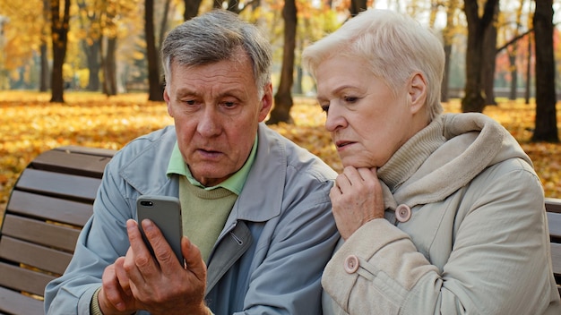 Una pareja caucásica casada molesta usa un teléfono inteligente sentado en un banco en el parque de otoño mira el teléfono de pantalla