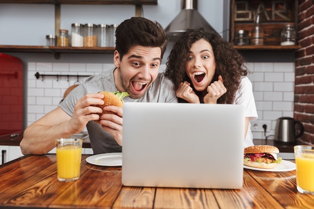 Pareja caucásica hombre y mujer mirando portátil en la mesa mientras come una hamburguesa en la cocina de casa