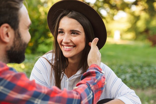 Pareja caucásica hombre y mujer vestidos con ropa casual sonriendo y mirando el uno al otro mientras descansa en el parque verde