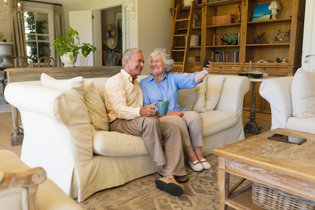 Una pareja caucásica mayor sentada en un sofá haciendo videollamadas usando un teléfono inteligente. concepto de retiro, jubilación y estilo de vida feliz para personas mayores.