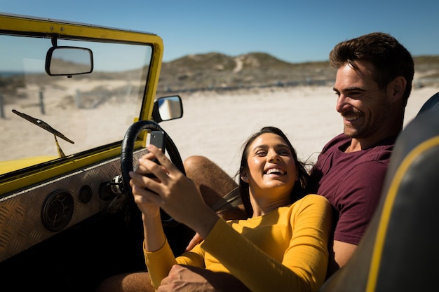 Una pareja caucásica tirada en un buggy de playa junto al mar usando un smartphone. descanso en la playa en un viaje por carretera de vacaciones de verano.