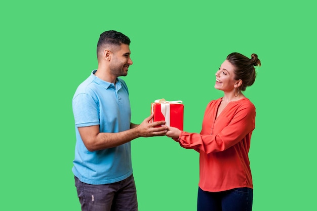 Pareja celebrando aniversario. Un hombre apuesto y positivo con ropa informal dando una caja con un regalo a una mujer encantadora, mirándose con amor el uno al otro. aislado sobre fondo verde, tiro de estudio interior