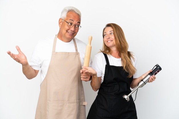 Pareja de chefs de mediana edad aislado sobre fondo blanco haciendo gestos sin importancia mientras levanta los hombros