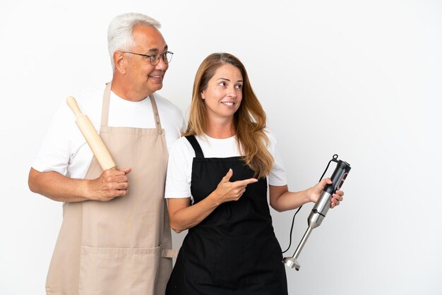 Pareja de chefs de mediana edad aislado sobre fondo blanco presentando una idea mientras mira sonriendo hacia