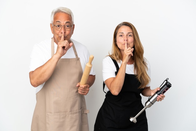 Pareja de chefs de mediana edad aislado sobre fondo blanco que muestra un signo de gesto de silencio poniendo el dedo en la boca