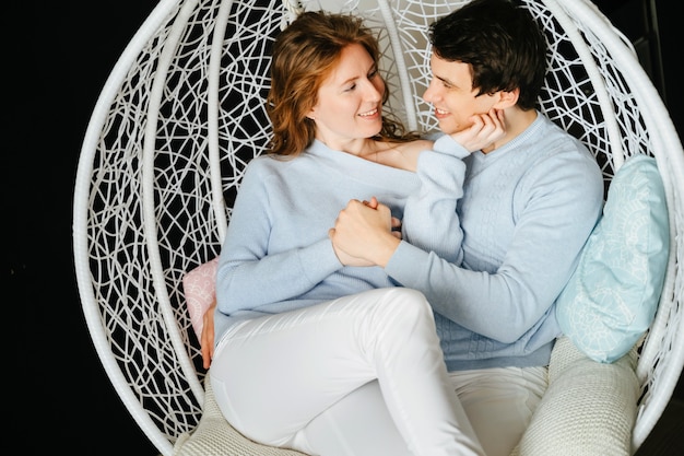 Pareja chica y chico abrazándose en una silla grande. Jersey blanco y azul.