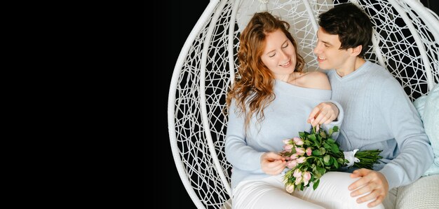 Pareja chica y chico abrazándose en una silla grande con un ramo de rosas. Jersey blanco y azul.