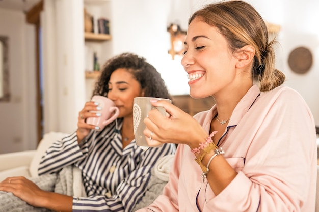 Pareja de chicas que cohabitan en pijama relajándose bajo una manta en el acogedor sofá beber una taza de té