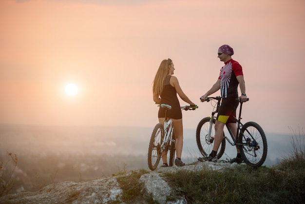 Pareja ciclista con bicicletas de montaña de pie sobre una roca, disfrutando del paisaje nocturno