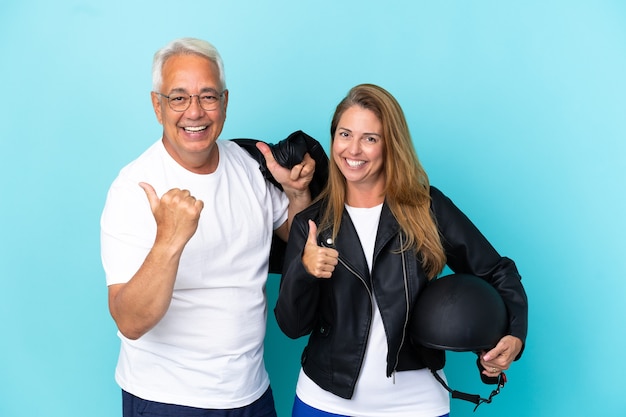 Pareja de ciclistas de mediana edad con un casco de motocicleta aislado sobre fondo azul dando un pulgar hacia arriba gesto con ambas manos y sonriendo