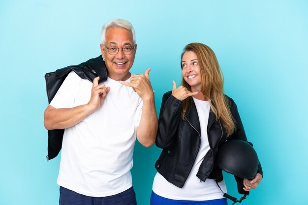 Pareja de ciclistas de mediana edad con un casco de motocicleta aislado sobre fondo azul haciendo gesto de teléfono. Llámame señal