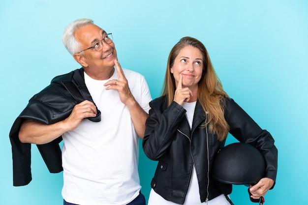 Pareja de ciclistas de mediana edad con un casco de motocicleta aislado sobre fondo azul pensando en una idea mientras mira hacia arriba