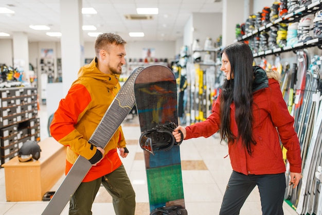 Pareja comprando esquí alpino y snowboard, compras en tienda de deportes.