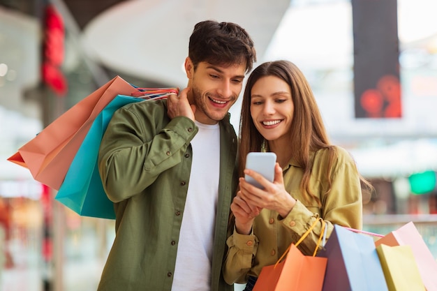 Pareja de compras usando la aplicación telefónica con bolsas de compras en el centro comercial