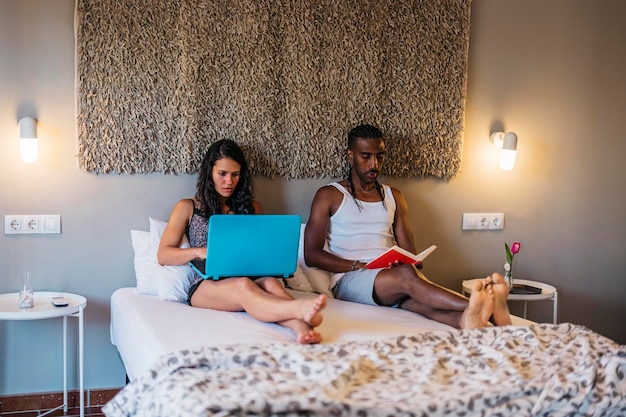 Una pareja comprometida sentada en una cama, ella con una laptop y él leyendo