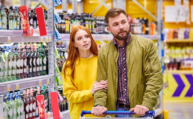 Pareja en el departamento de alcohol en el supermercado, hacer una elección, mirar los estantes con botellas, en el pasillo