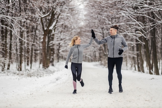 Pareja deportiva corriendo juntos en un día nevado de invierno en el bosque. Se dan cinco el uno al otro. Fitness al aire libre, fitness de invierno, hábitos saludables.