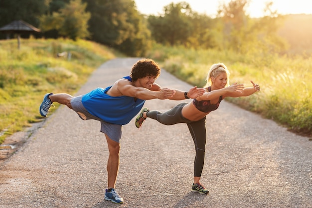 Foto pareja deportiva haciendo ejercicio de equilibrio sobre un pie en la carretera en la naturaleza. longitud total, día soleado de verano.