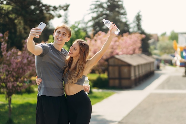 Pareja deportiva tomando selfie en el teléfono inteligente durante el descanso en el entrenamiento al aire libre posando en la cámara y sonriendo