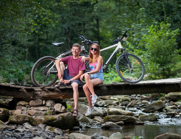 Pareja descansando en el bosque en el puente del río cerca de bicicletas