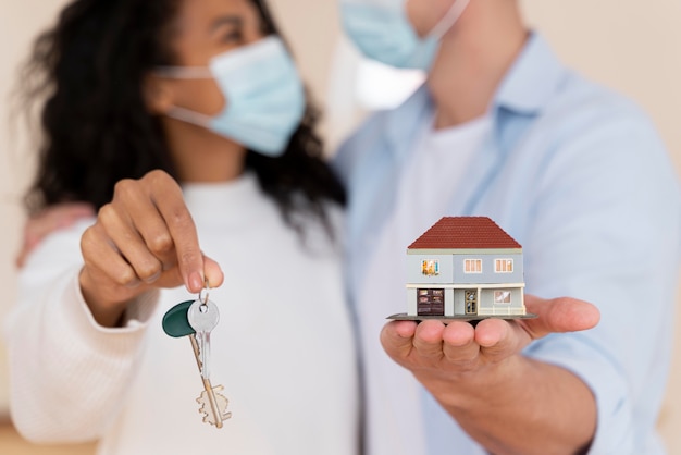 Pareja desenfocada con máscaras médicas sosteniendo nuevas llaves de casa y casa en miniatura