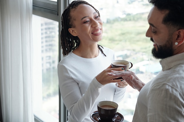 Pareja devirse positiva de pie en la ventana del apartamento y disfrutando de un delicioso café matutino