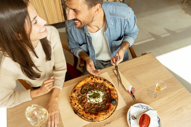 Una pareja disfrutando de una deliciosa comida en un acogedor restaurante mientras comparten una deliziosa pizza y un vaso de vino blanco