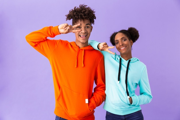 Pareja divertida en ropa colorida sonriendo y mostrando el signo de la paz, aislado sobre la pared violeta