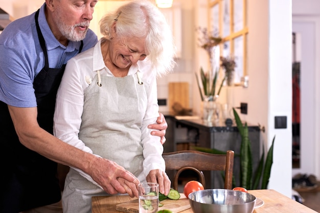 Pareja divirtiéndose en la cocina con comida sana, cocinando comida en casa, preparando el almuerzo con verduras frescas bio, tallando o cortando verduras, el hombre ayuda a su esposa, vistiendo delantal