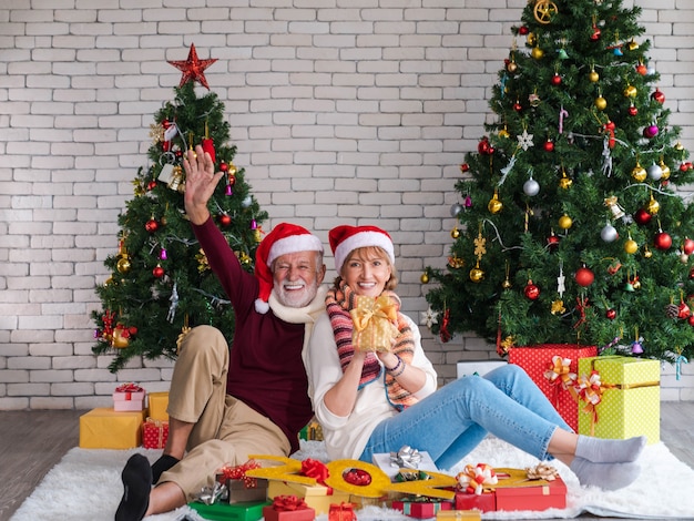 Pareja de dos adultos mayores felices y alegres con gorro de Papá Noel celebrando año nuevo, nueva vida y Navidad en casa con árbol de Navidad decorado en sala de estar con regalos y número 2022. adiós.