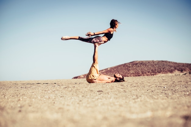 Pareja de dos adultos y personas juntas en la playa entrenando juntos en la arena haciendo posiciones de yoga con colinas al fondo