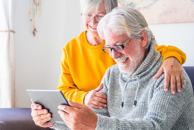 Pareja de dos ancianos alegres y felices maduros y ancianos usando tableta y divirtiéndose juntos sentados en el sofá en casa. Jubilados de belleza que utilizan internet y navegan por la red