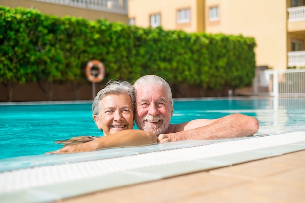 Pareja de dos ancianos felices divirtiéndose y disfrutando juntos en la piscina sonriendo y mirando a la cámara. Gente feliz disfrutando del verano al aire libre en el agua