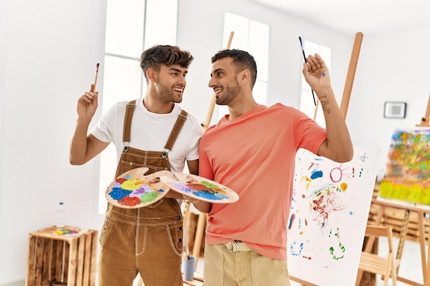 Pareja de dos hombres hispanos sonriendo confiados sosteniendo pinceles y paleta en el estudio de arte