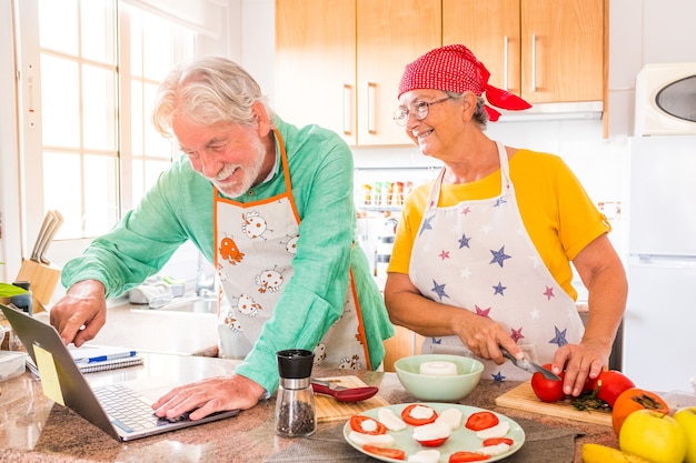 Pareja de dos personas mayores felices cocinando juntos en la cocina aprendiendo y siguiendo un video tutorial de cocinar en la computadora portátil: preparar alimentos saludables juntos