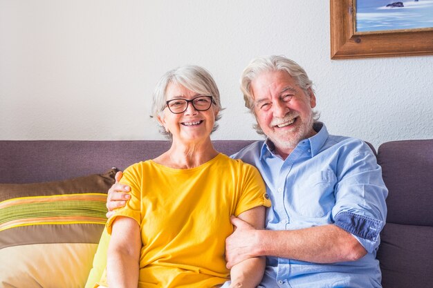 Pareja de dos personas mayores felices o personas maduras sonriendo y mirando a la cámara sentado en el sofá en casa - retrato de jubilados sentados en el sofá en el interior