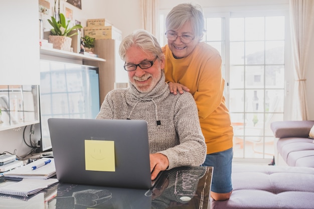 Pareja de dos personas mayores felices trabajando y usando la computadora portátil juntos en casa sonriendo y divirtiéndose juntos - estilo de vida de bloqueo